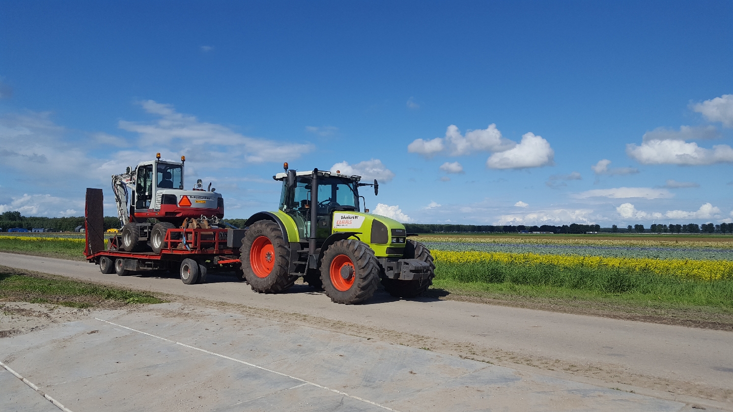 Groene Claas-trekker met Schuchard BV-sticker trekt een dieplader met een kraan langs de bloembollenvelden
