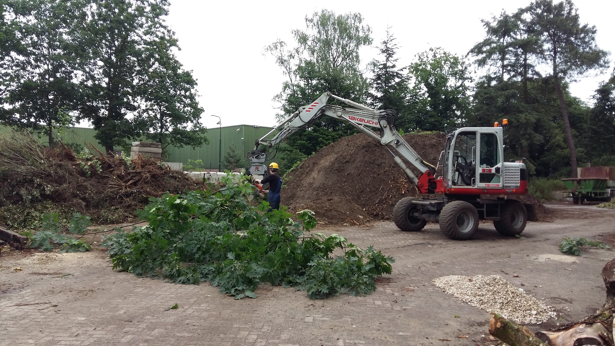 Kraan en medewerker verwerken groenafval naast een berg grond